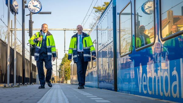 Twee mannelijke machinisten in uniform lopen over een perron. Een blauwe Arriva-trein staat op het spoor.