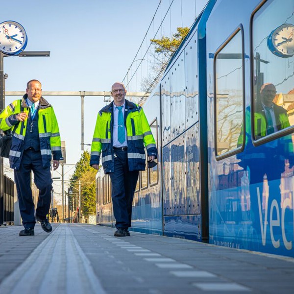Twee mannelijke machinisten in uniform lopen over een perron. Een blauwe Arriva-trein staat op het spoor.