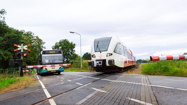 Een groene bus van Arriva staat te wachten voor een spoorovergang in Zutphen waar een rode trein van Arriva passeert.