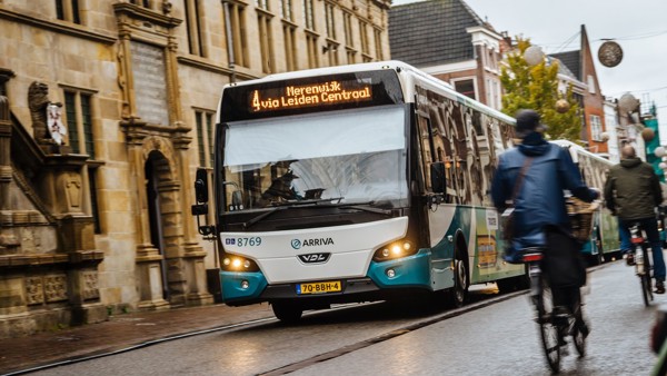 Blauw-witte Arriva-bus rijdt door het centrum van Leiden langs een statig gebouw, met fietsers op de weg.