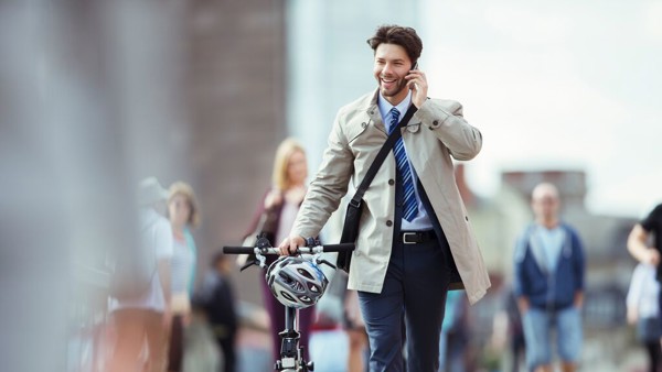 Een bellende, lachende man in business-casual outfit loopt met vouwfiets en helm aan de hand door een stad.