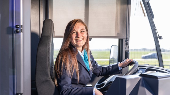 Vrouwelijke buschauffeur met lang haar en in uniform zit lachend achter het stuur van haar Arriva-bus.