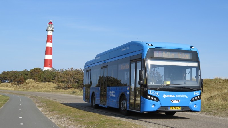 Een blauwe Arriva-bus met daarop Ballum - Hollum rijdt door de duinen met een rood-witte vuurtoren op de achtergrond.