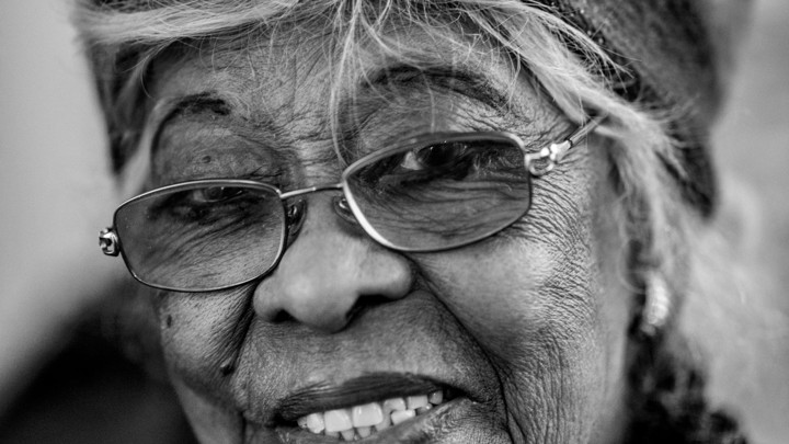 Een knappe, oude, lachende vrouw met een bril op en haarband in over haar krullende haar lacht naar de camera.