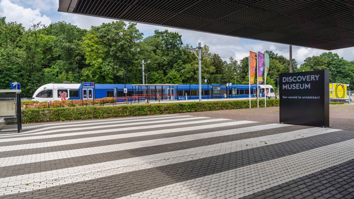 Blauwwitte trein van Arriva staat aan perron van station Kerkrade, op de voorgrond een zebrapad en een bordje met 'Discovery Museum'.