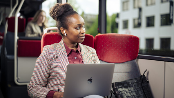 Een vrouw zit in de bus in pak met oordopjes in en laptop op schoot terwijl ze uit het raam kijkt.