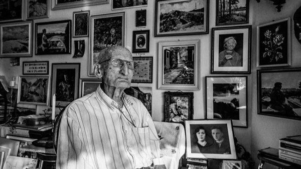 Een zwart-witfoto met daarop een heel oude man in overhemd die in de verte staart, de muur achter hem is volgehangen met foto's.